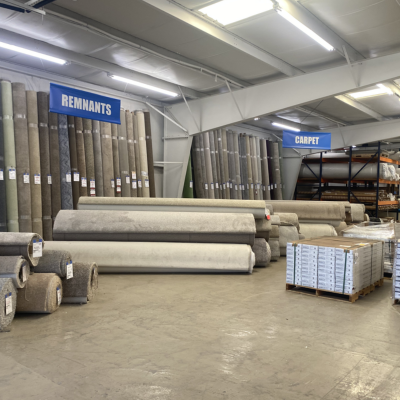 Bright carpet warehouse with vertical rolls in beige, tan, and gray lining walls beneath blue Remnants and Carpet signs.